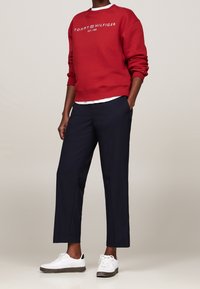 Rode sweatshirt met witte "TOMMY HILFIGER" opdruk, gecombineerd met marineblauwe broek. Model draagt witte sneakers met zwarte accenten.