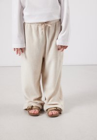Enfant portant un pantalon beige ample avec une taille à cordon, une chemise blanche à manches longues et des sandales marron à bouts ouverts, debout sur un sol uni.