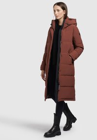 Lange, bruine gewatteerde jas met een capuchon, ritssluiting, zakken en zwarte accenten. Draagbaar over een zwarte top, gecombineerd met zwarte enkellaarzen.