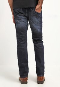 Pantalones de mezclilla azul oscuro con corte ajustado, que presentan bolsillos traseros inclinados y un sutil desteñido. Combinados con zapatos de cuero marrón.