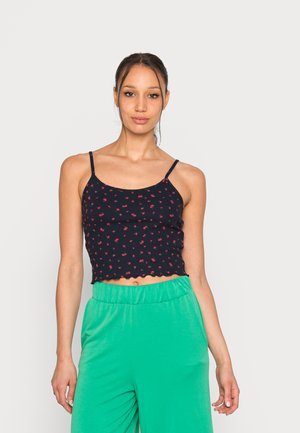 Top cropped azul-marinho com pequeno padrão floral vermelho, bainha em forma de ondas e alças finas ajustáveis, combinado com calças verdes largas.