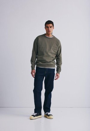 Olive green sweatshirt z okrągłym dekoltem, noszony na białej koszuli. Połączony z ciemnoniebieskimi dżinsami i czarnymi trampkami z żółtymi podeszwami.