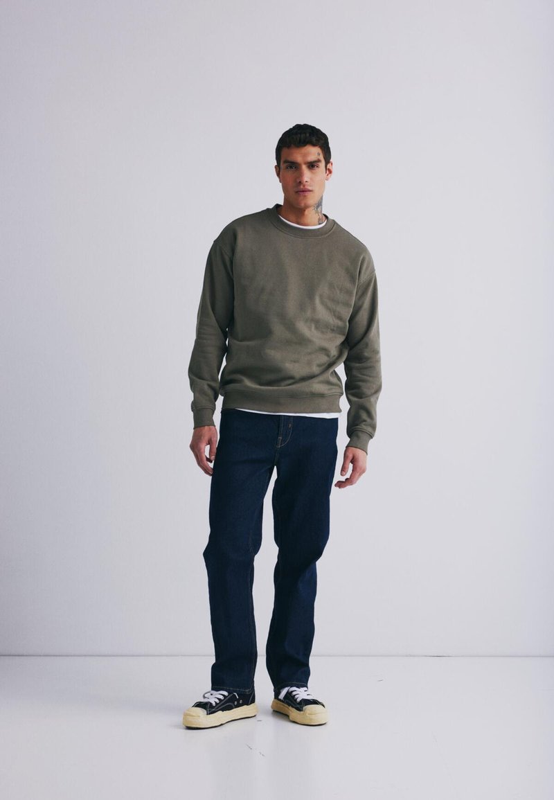 Alyvuogių žalias megztinis su apvaliu kaklu, uždėtas ant balto marškinio. Sutapatintas su tamsiai mėlynomis džinsomis ir juodais sportbačiais su geltonais padais.