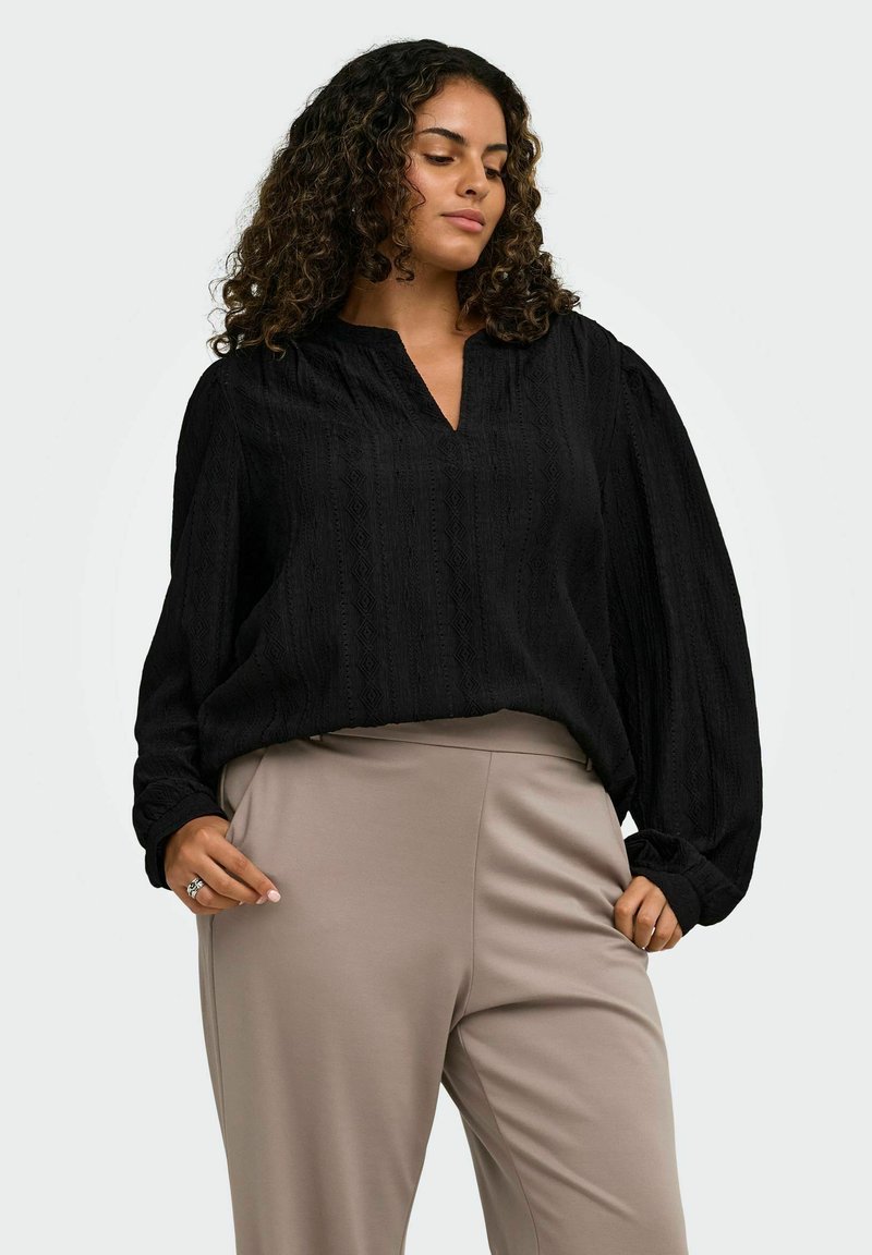 Femme aux cheveux bouclés portant un chemisier noir à manches longues et un pantalon beige, debout devant un fond clair uni.