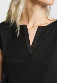 Svart kortärmad topp med V-ringning och subtil knäppning, tillverkad av ett mjukt tyg med en lätt struktur.