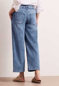 Persona que lleva jeans de pierna recta de color azul claro con bolsillos traseros y zapatos slip-on color beige, de pie con las manos en los bolsillos contra un fondo liso.