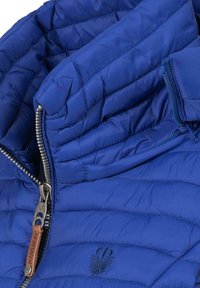 Blauwe pufferjack met een hoge kraag, horizontaal gestikte patroon, zilveren ritsluiting en een decoratief logo nabij de zoom.