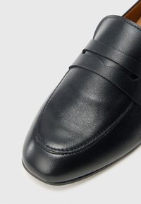 Schwarze Leder-Loafers mit glatter Textur, abgerundeter Spitze und dekorativem Riemen über dem Spann mit Ziernaht.