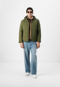 Olivegrüne Kapuzenjacke mit Reißverschluss, über einem braunen Hemd getragen. Kombiniert mit hellblauen weiten Jeans und weißen Sneakers. Runde Sonnenbrille.
