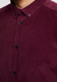 Dunkelrotes Cordhemd mit Button-Down-Kragen, schwarzen Knöpfen und gerippter Textur. Nahaufnahme zeigt die Details des Stoffes.