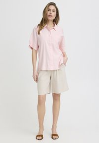 Femme debout portant une chemise à manches courtes rose clair à boutons, un short beige à hauteur du genou et des talons ouverts marron sur un fond blanc uni.