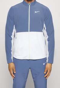 Nike jacket in blue and white, featuring a zip-front design, stand collar, and textured mesh accents. Smooth fabric with logo detail.
