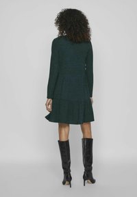 Robe vert foncé avec un design à volants, manches longues, texture côtelée et taille cintrée ; associée à de grandes bottes noires à talons.