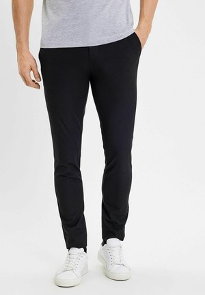 Man draagt slim zwarte broek met voorzakken en witte sneakers, staand tegen een effen lichte achtergrond.