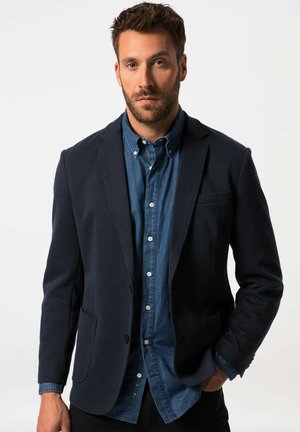 Homme aux cheveux bruns courts et à la barbe, portant un blazer sombre sur une chemise bleue boutonnée, debout avec une main dans la poche.