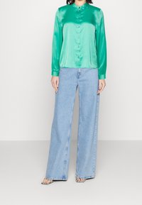 Camicia verde in satin a maniche lunghe con chiusura a bottoni, abbinata a jeans a gamba larga blu chiaro e sandali con stampa animale. Sfondo semplice.