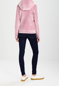 Person i en ljusrosa huvad jacka, mörka skinny jeans och vita sneakers med gummisnäckor, stående mot en enkel, ljus bakgrund.