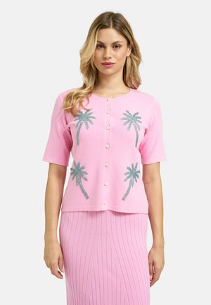 Frau trägt eine rosa kurzärmelige, geknöpfte Strickjacke mit grünen Palmen-Motiven, kombiniert mit einem rosa gerippten Rock.