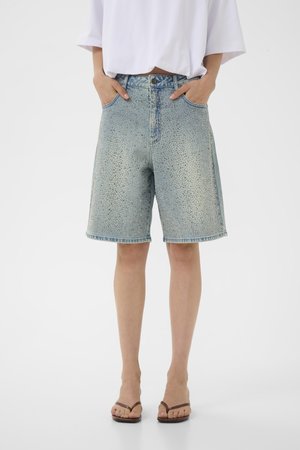Personne portant un short en jean bleu clair avec des strass, un t-shirt blanc ample et des sandales plates marron, mains dans les poches, debout sur fond blanc.