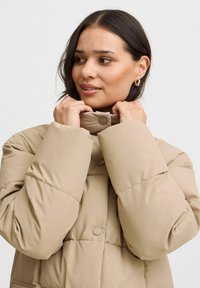 Chaqueta acolchada beige con cuello alto, cierres de botones y mangas amplias, fabricada con un material ligero y suave al tacto.
