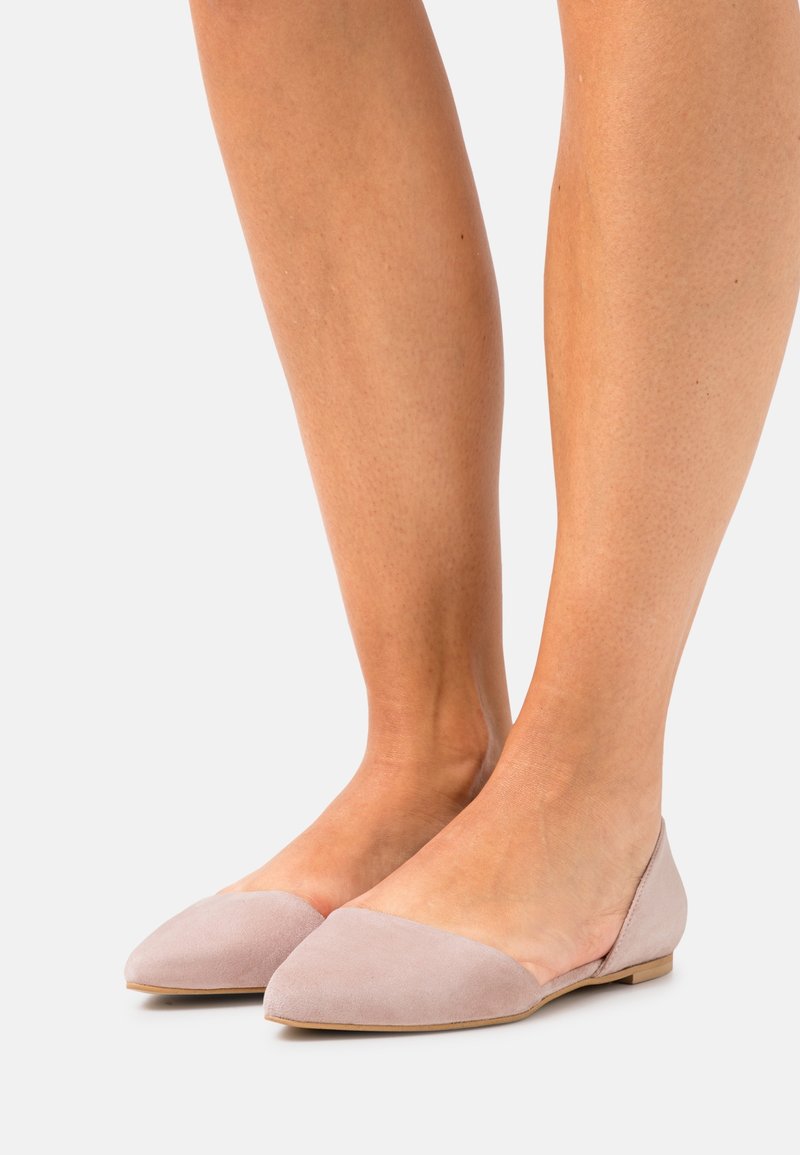 Personne portant des chaussures plates rose clair à bout pointu, debout les pieds rapprochés sur un fond uni.