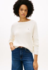 Λευκό πλεκτό πουλόβερ με ριμπ ύφανση, φαρδύ ντεκολτέ και μακριά μανίκια, συνδυασμένο με ψηλόμεσο μπλε τζιν. Μικρή λεπτομέρεια λογότυπου στο πουλόβερ.