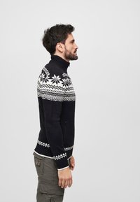 Suéter negro de punto con cuello alto, que presenta patrones de copos de nieve blancos y geométricos en el pecho y el dobladillo. Detalle de cremallera en el lateral.