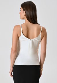 Witte satijnen camisole met dunne verstelbare bandjes, met een lage rug, kleine decoratieve knoop en een gladde textuur. Gecombineerd met een zwarte onderkant.