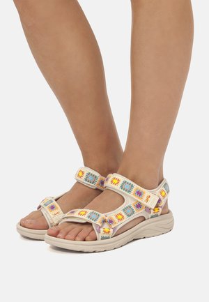 Füße, die beige Sandalen mit bunten, gewebten Quadratmustern und verstellbaren Riemen tragen, vor einem hellgrauen Hintergrund.