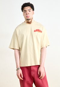 Beige ylisuuri t-paita, jossa on "HAWAII" punaisella ja keltaisella tekstillä, yhdistettynä punaisiin housuihin. Kangas näyttää sileältä ja rentolta.