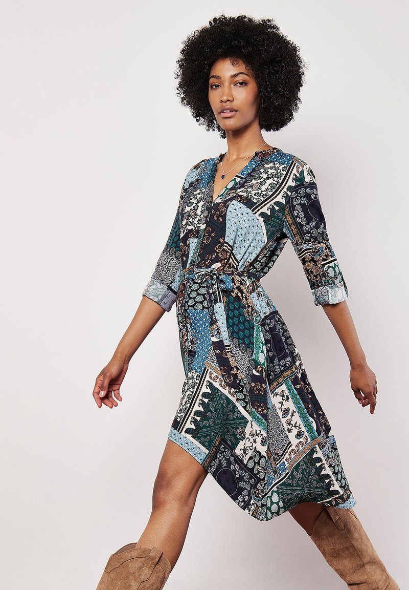 Šaty s více vzory, které kombinují tmavě modrou, zelenou a krémovou barvu. Jsou vyrobeny z lehkého materiálu, mají zavinovací design a 3/4 rukávy.