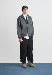 Grauer Sweatshirt mit Reißverschluss und zwei Vordertaschen, darüber ein hellrosa Hemd und eine schwarze Krawatte getragen. Kombiniert mit schwarzen Cargo-Hosen und beigen Stiefeln.