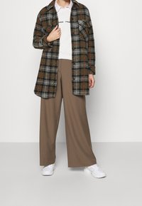 Camisa de franela a cuadros en gris, verde y naranja con botones y dos bolsillos en el pecho, combinada con pantalones marrones holgados y zapatillas blancas.