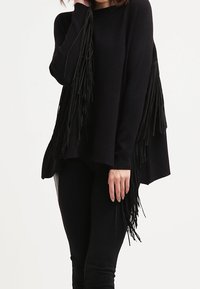 Kvinna som bär en svart långärmad topp med fransdetaljer på ärmar och sidor, tillsammans med svarta tajta byxor.