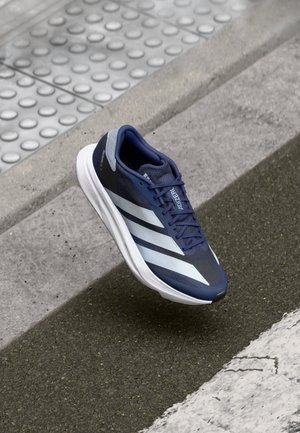Blauer Adidas-Laufschuh mit weißen Streifen schwebt über einem strukturierten Bürgersteig und Straßenbordstein.