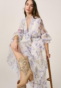 Mujer sentada en taburete con vestido blanco translúcido con estampado floral morado y botas de vaquero beige con recortes y flecos.
