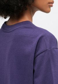 T-shirt di cotone viola con scollatura rotonda e maniche corte, caratterizzato da una texture liscia e un sottile dettaglio di cucitura sulla spalla.