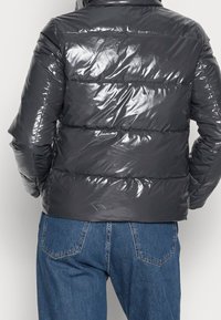 Dopasowana kurtka puchowa w błyszczącym ciemnoszarym kolorze z poziomymi przeszyciami, wysokim kołnierzem i materiałem odpornym na wodę. Pod spodem widoczne dżinsy.