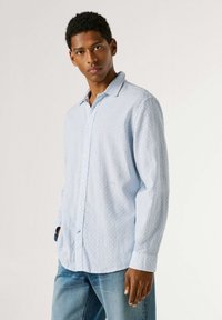 Young man wearing light blue patterned long-sleeve shirt and blue jeans, standing against plain white background.