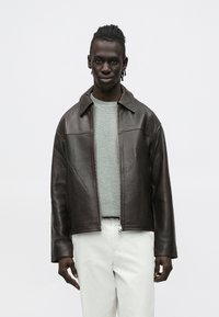 Brūna ādas jaka ar apkakli, priekšējo rāvējslēdzēju un divām sānu kabatām, valkāta pār pelēku džemperi un baltām biksēm. Gluda tekstūra.