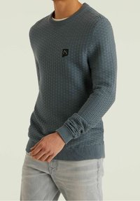 Sweater in textured blue fabric with a ribbed collar and cuffs. Features a small black logo patch on the left chest. Casual fit.