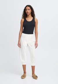 Un débardeur noir à côtes associé à un pantalon cropped blanc, ample et à taille smockée. Le modèle porte des mocassins en cuir souple marron clair.