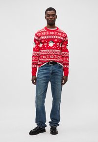 Roter Weihnachtspullover mit Schneeflocken- und Schneemann-Mustern, getragen über blauen Jeans. Der Pullover hat gerippte Bündchen und einen Kragen, eine feste Textur und polierte schwarze Schuhe.