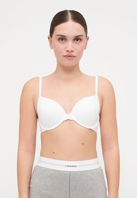 Hvit polstret bh med justerbare stropper og sømløst design. Materialet ser glatt ut med en litt skinnende tekstur. Grå midjebånd synlig under.