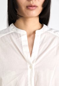 White collared blouse made of light fabric, featuring a buttoned placket, subtle texture, and a relaxed fit, viewed from the chest upwards.
