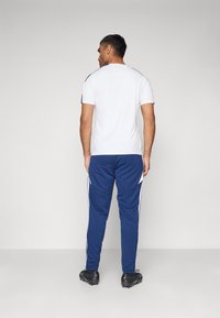 Weißes Sport-T-Shirt mit schwarzen Schulterakzenten, kombiniert mit blauen Jogginghosen mit weißen Seitenstreifen und strukturiertem Stoff. Schwarze Schuhe sind sichtbar.