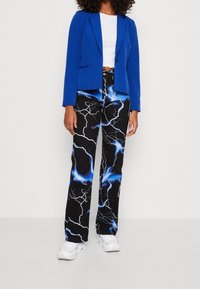 Blå kavaj med en figurnära design, bärs över en vit crop top. Svarta byxor med ett blått blixtmönster, kombinerade med vita sneakers.