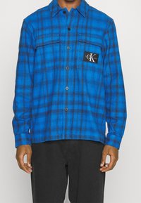 Blue plaid flannel shirt featuring black accents, button closure, and a patch with the logo. Long sleeves with foldable cuffs.