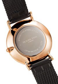 Reloj de caja redonda en oro rosa con fondo de acero inoxidable. Correa de malla negra. Presenta texto grabado en la parte trasera, indicando la marca y el tipo de movimiento.