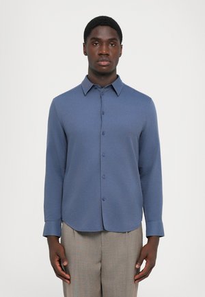 Blauwe langarmsgemouwen shirt met een knoopsluiting aan de voorkant, puntige kraag en afgeronde zoom; gemaakt van een gestructureerde stof. Draagt men met lichtgrijze broeken.